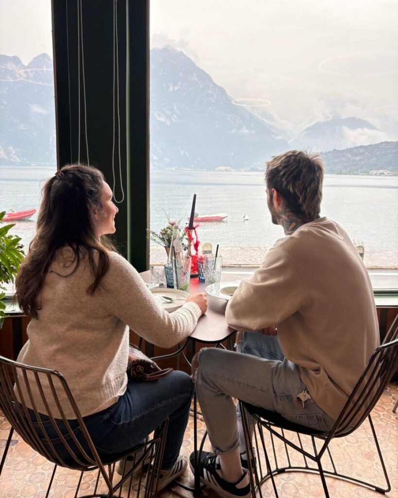 Couple enjoying scenic lake view from restaurant. Mountains, boats, idyllic scene. Travel photography, vacation vibes.