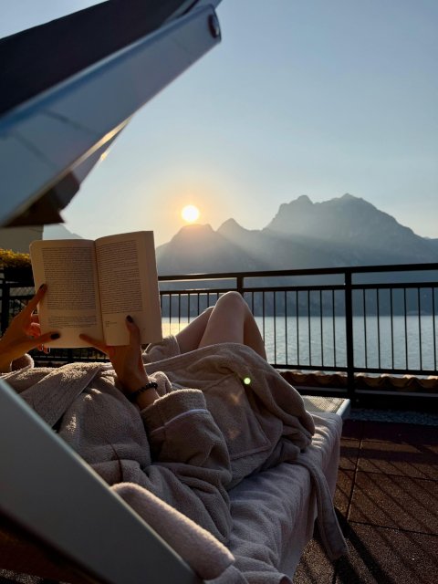 Woman reading book on lounge chair, mountain view, lake, sunset. Relaxation and vacation lifestyle concept.