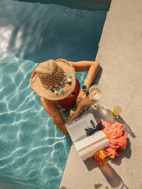 Woman in straw hat relaxing in a swimming pool with a book, sunglasses, and a cocktail on a sunny summer vacation day.