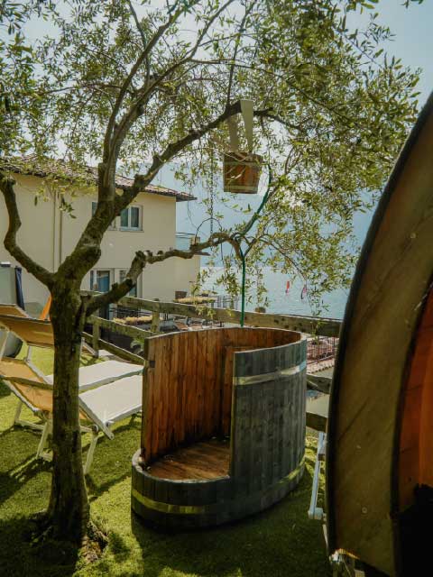 Outdoor wooden barrel shower in a garden with olive trees, lounge chairs, and a scenic lake view near a house.