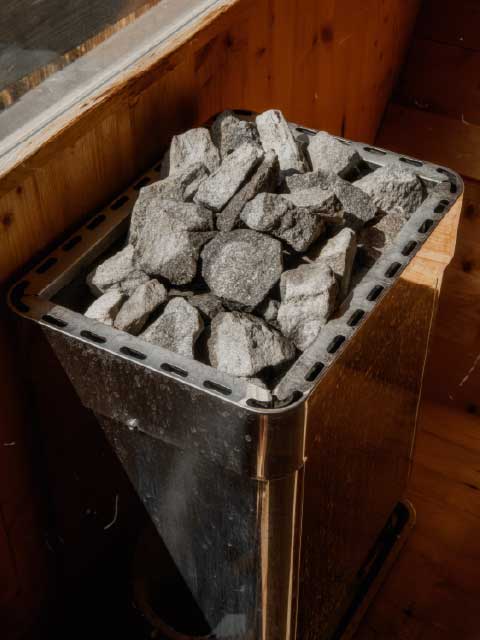 Close-up of a stainless steel sauna heater filled with natural grey volcanic stones in a rustic wooden sauna room.